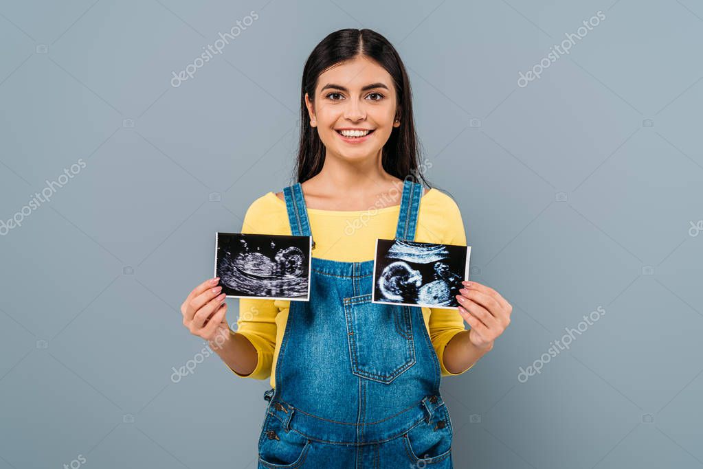 Smiling pregnant pretty girl holding fetal ultrasound images isolated on grey