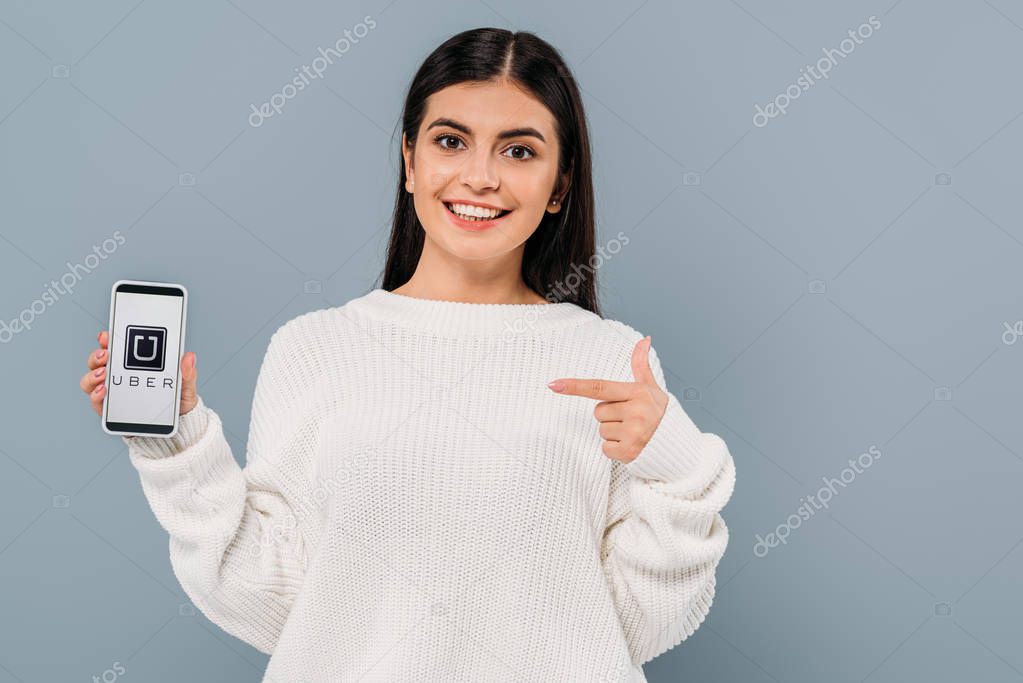 KYIV, UKRAINE - SEPTEMBER 20, 2019: smiling pretty brunette girl in white sweater pointing with finger at smartphone with uber app isolated on grey