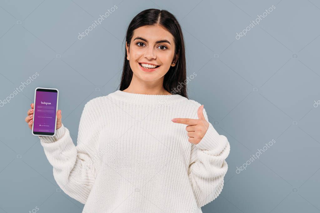 KYIV, UKRAINE - SEPTEMBER 20, 2019: smiling pretty brunette girl in white sweater pointing with finger at smartphone with instagram app isolated on grey