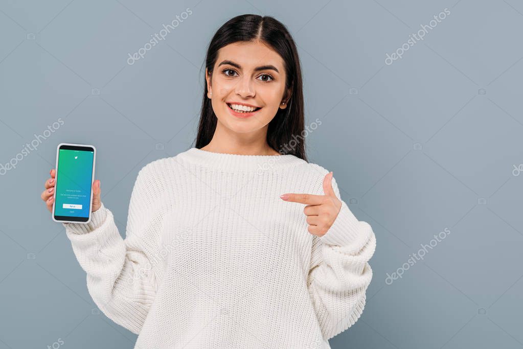 KYIV, UKRAINE - SEPTEMBER 20, 2019: smiling pretty brunette girl in white sweater pointing with finger at smartphone with twitter app isolated on grey