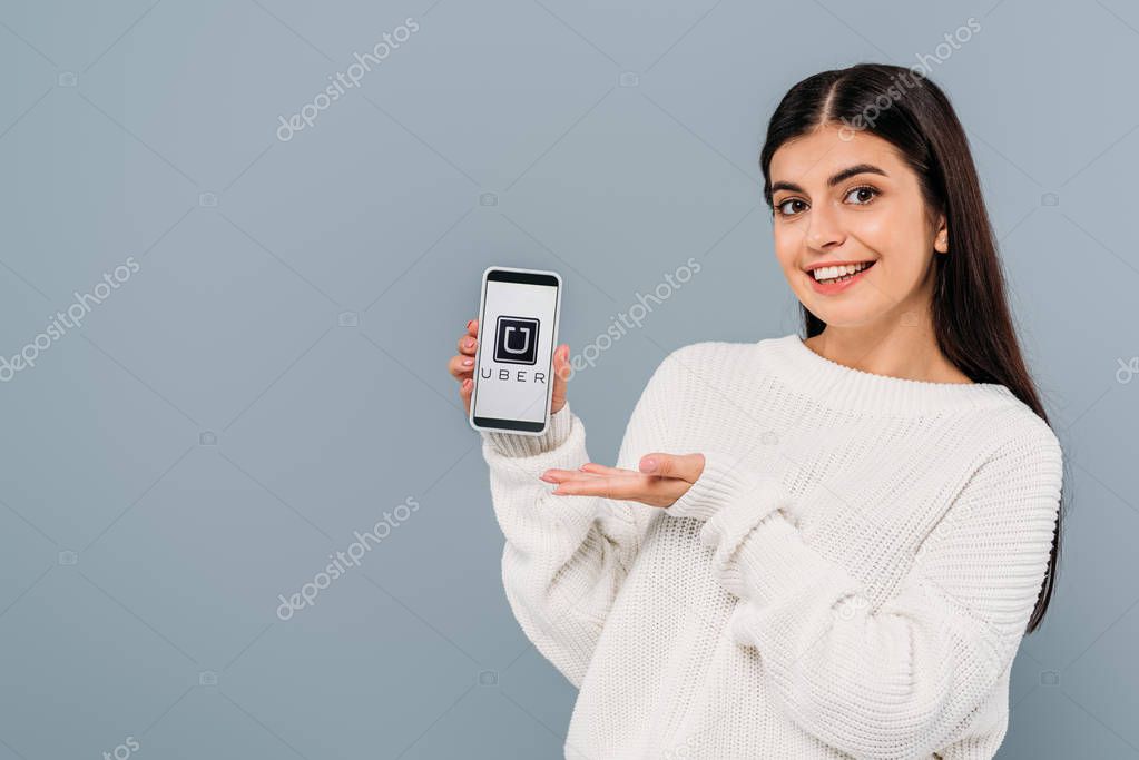 KYIV, UKRAINE - SEPTEMBER 20, 2019: smiling pretty brunette girl in white sweater holding smartphone with uber isolated on grey