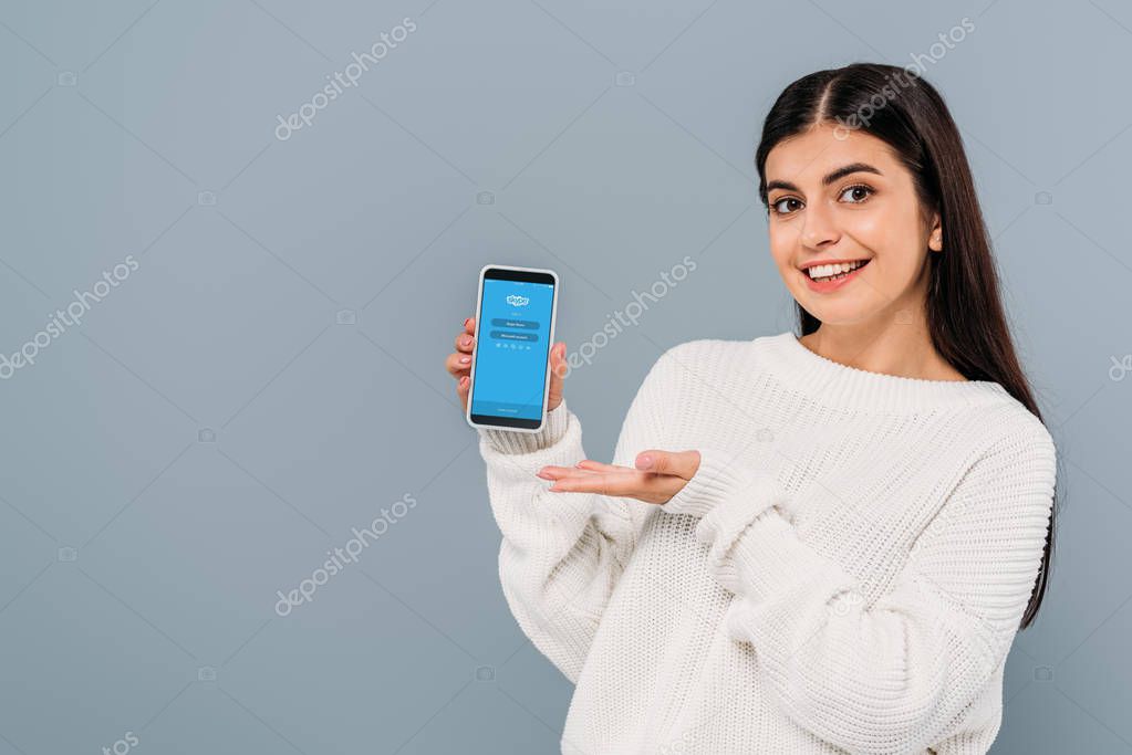 KYIV, UKRAINE - SEPTEMBER 20, 2019: smiling pretty brunette girl in white sweater holding smartphone with skype app isolated on grey