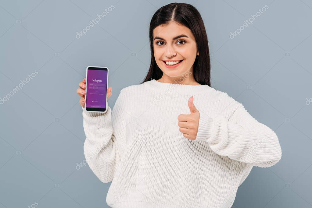 Smiling pretty girl in white sweater showing smartphone with instagram app and thumb up isolated on grey