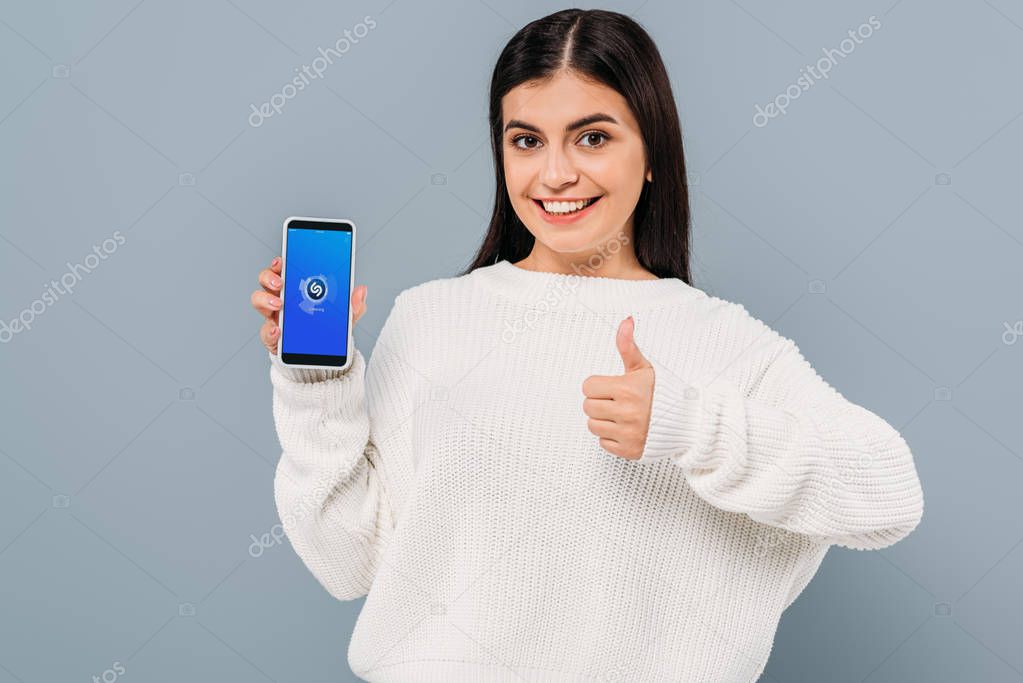 Smiling pretty girl in white sweater showing smartphone with shazam app and thumb up isolated on grey