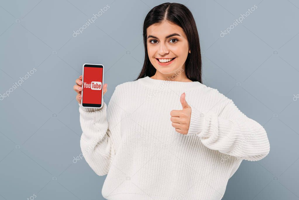 Smiling pretty girl in white sweater showing smartphone with youtube app and thumb up isolated on grey