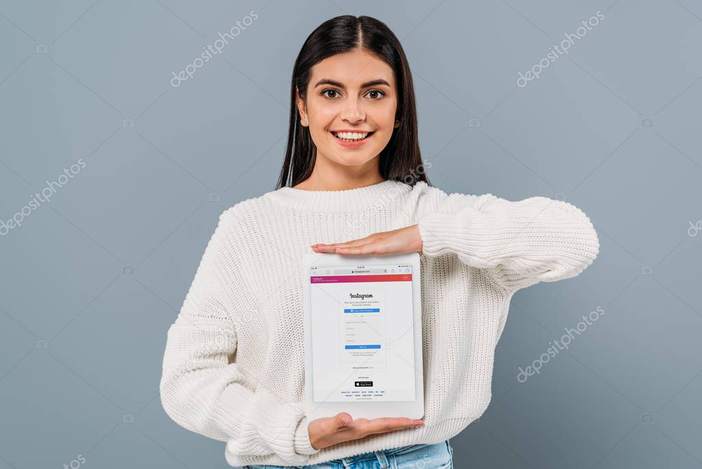 KYIV, UKRAINE - SEPTEMBER 20, 2019: smiling pretty brunette girl in white sweater holding digital tablet with instagram app isolated on grey
