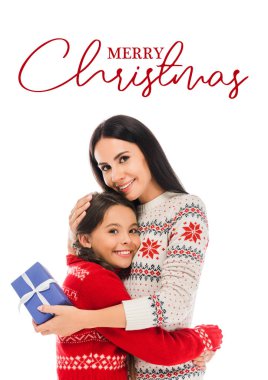 cheerful mother hugging daughter and holding preset isolated on white with merry Christmas lettering clipart