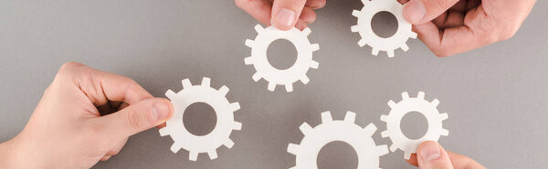 обрезанный вид людей, держащих белые шестерни на сером фоне, панорамный снимок
