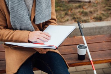 Cropped view of blind man reading book with braille font beside walking stick and thermo mug on bench clipart