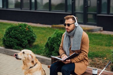 Kulaklıklı kör adam rehber köpeğin yanında kitap tutuyor. 