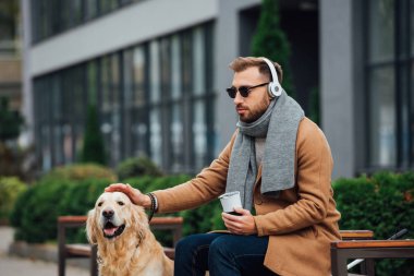 Blind man in headphones holding thermo mug and stroking guide dog in park clipart