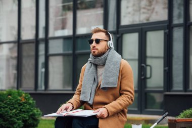 Blind man in headphones reading book on bench in park clipart