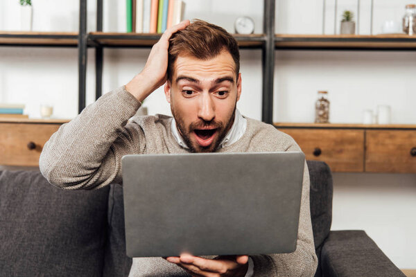 Шокированный человек смотрит на ноутбук на диване в гостиной
