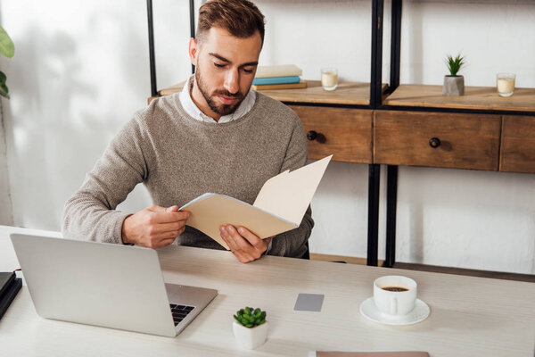 Man holding paper folder beside laptop and coffee on table