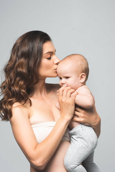 portrait of attractive mother kissing baby boy, isolated on grey