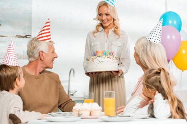 Güzel bir kadın, ailenin yanında mutfak masasında oturan yaşlı adama doğum günü pastasını sunuyor.