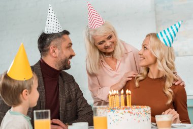Gülümseyen yaşlı kadın mutlu bir kadının omuzlarına dokunuyor doğum günü pastasının yanında ailesiyle oturuyor.
