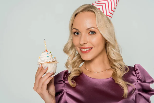 誕生日のカップケーキを持ってパーティーキャップで魅力的で陽気な女性とグレーで隔離されたカメラを見て — ストック写真