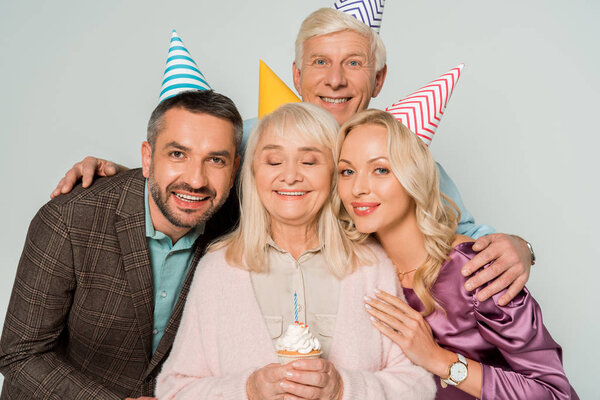 happy senior woman holding birthday cupcake near husband and adult daughter and son isolated on grey
