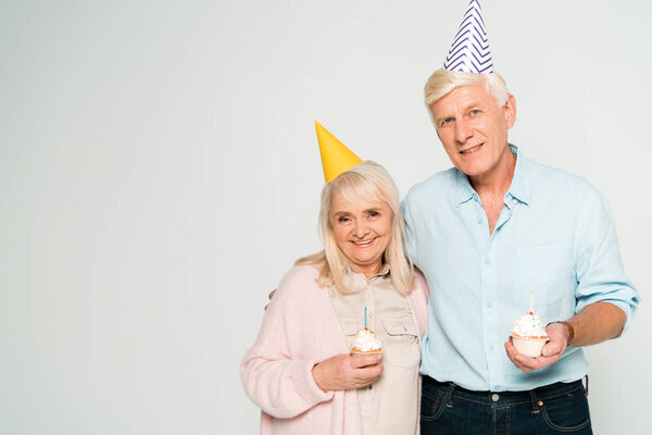 счастливые старший муж и жена с кексами на день рождения и улыбаясь в камеру изолированы на серый
