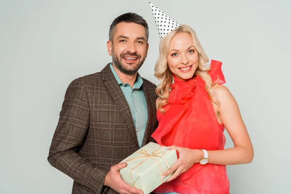 happy husband and wife smiling at camera while holding gift box isolated on grey