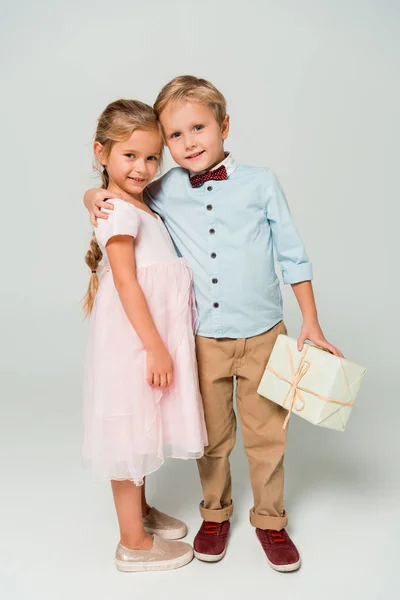 可爱的男孩拿着礼品盒 抱着灰色背景的可爱的妹妹 — 图库照片
