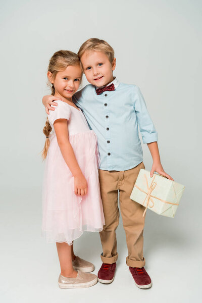 cute boy holding gift box and embracing adorable sister on grey background