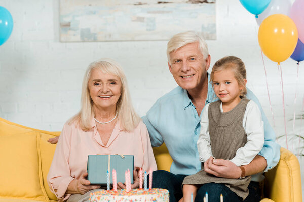 счастливая пожилая женщина держит подарочную коробку, сидя на диване рядом муж и внучка
