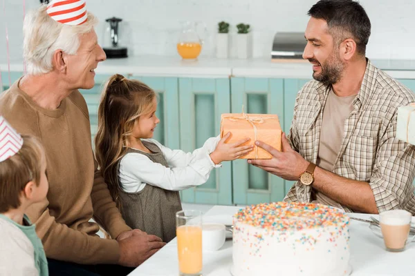 幸せな男は娘からギフトボックスを取る家族と一緒に誕生日ケーキの近くに座っている間 — ストック写真