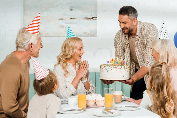 Glimlachende Man Met Verjaardagstaart Met Kaarsen Buurt Van Gelukkig Gezin — Stockfoto