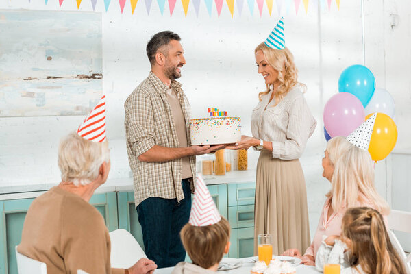 Улыбающийся мужчина представляет торт на день рождения счастливой жене, в то время как вся семья сидит за кухонным столом
