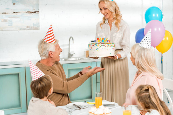 Улыбающаяся женщина представляет торт на день рождения старшему мужчине, сидящему за кухонным столом рядом с семьей
