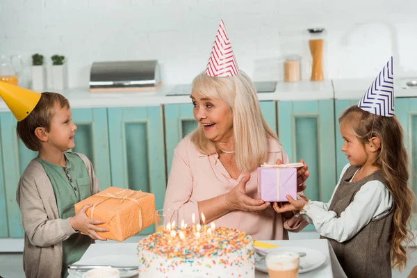 Adorable Kids Presenting Gift Boxes Happy Granny Sitting Birthday Cake — Stock Photo, Image