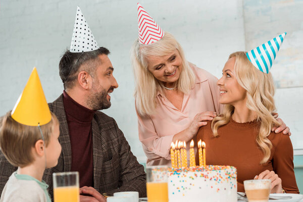 Улыбающаяся пожилая женщина трогает плечи счастливой женщины, сидящей рядом с праздничным тортом с семьей
