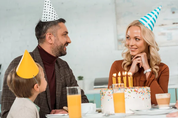 happy husband and wife looking at each other while sitting at kitchen table with son near birthday cake