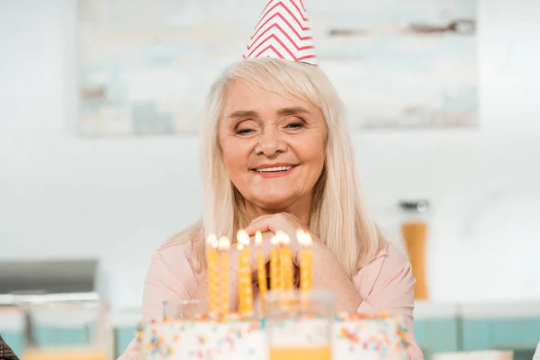 戴着派对帽的快乐的老年妇女看着点着蜡烛的生日蛋糕 — 图库照片