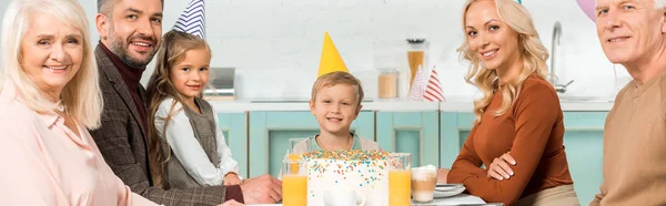 穿着派对帽坐在生日蛋糕旁边看着相机的快乐家庭的全景照片 — 图库照片
