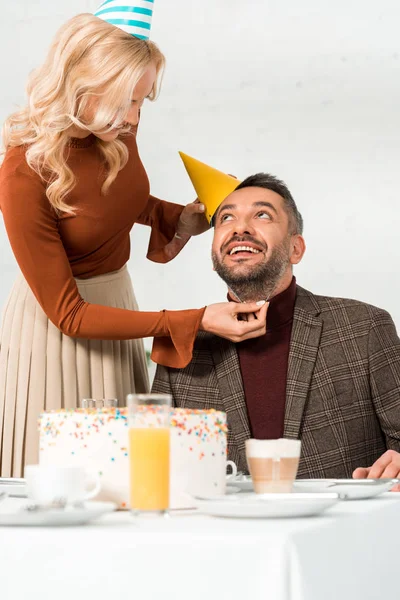 Mujer Sonriente Poniendo Gorra Fiesta Marido Feliz Sentado Mesa Con — Foto de Stock