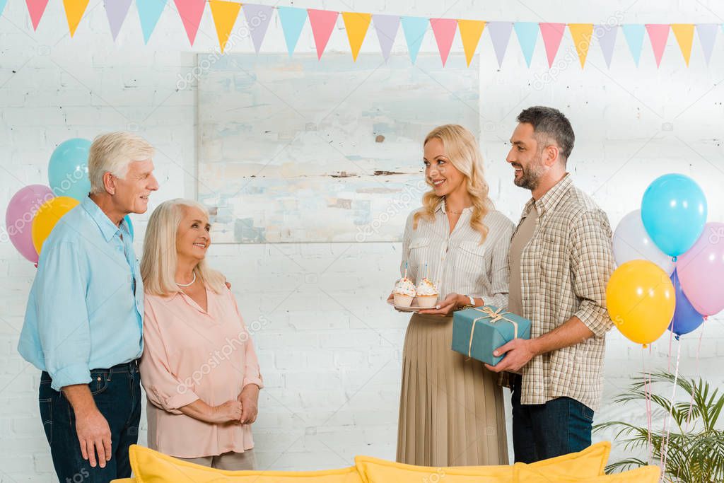 smiling man and woman holding birthday cupcakes and gift box while looking at senior parents