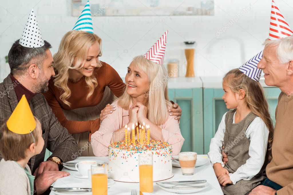 smiling woman touching shoulders of happy senior woman while all family sitting near birthday cake