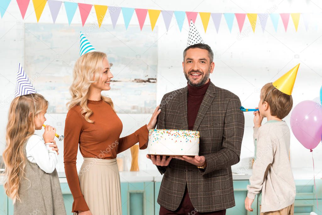 happy man looking at camera while holding birthday cake near wife and kids
