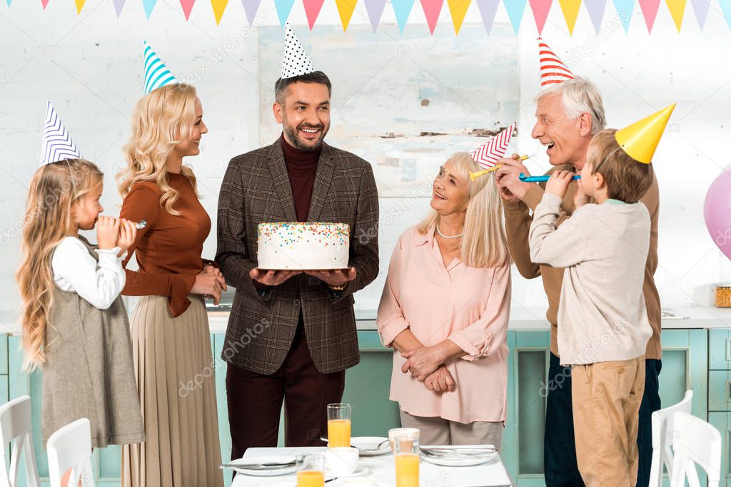 happy man holding birthday cake near cheerful family in party caps