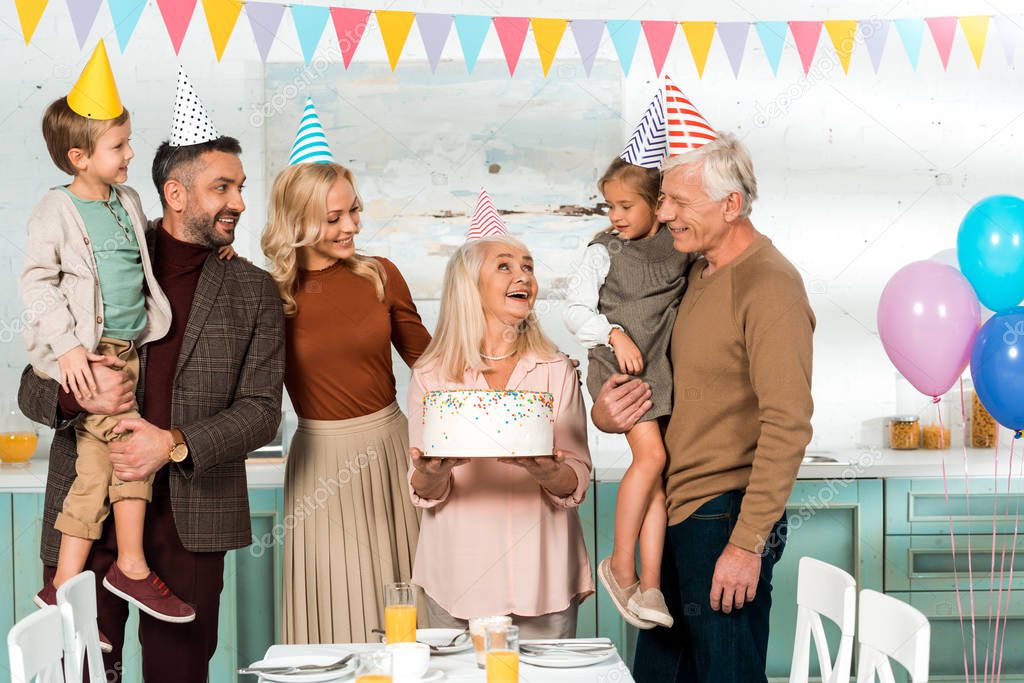 happy senior woman holding birthday cake near cheerful family in party hats