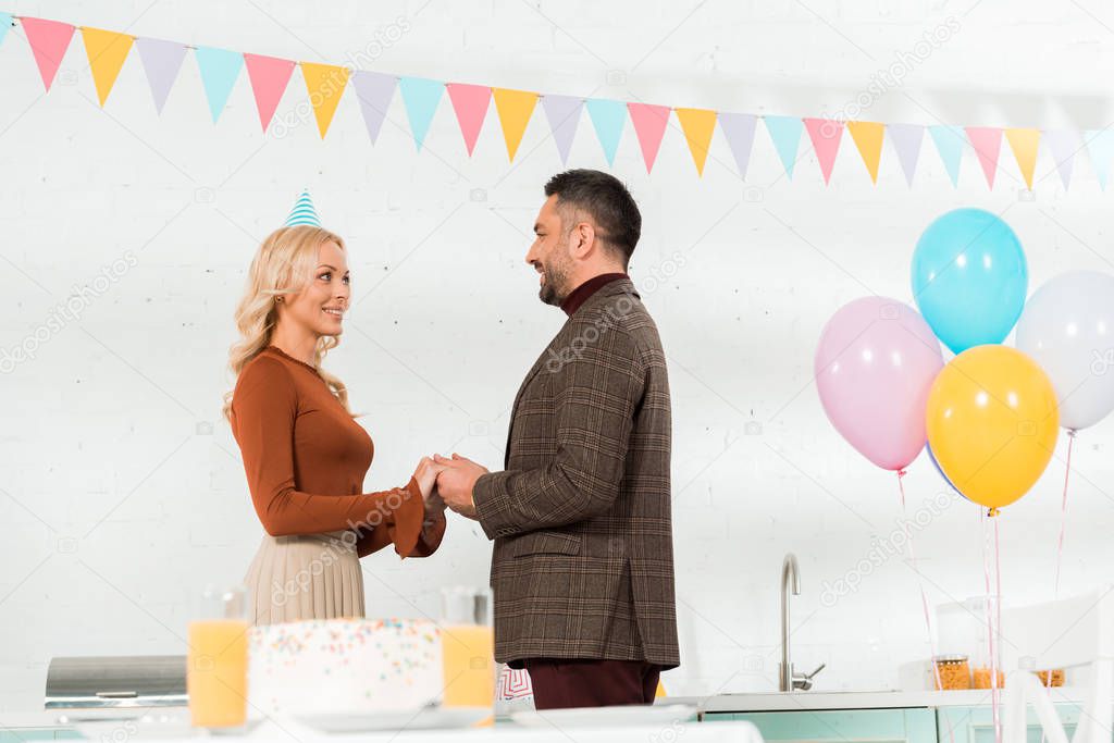 happy woman holding hands with husband near birthday cake and colorful balloons