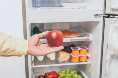 Kırpılmış görüntüde, kırmızı elmayı yiyecek dolu buzdolabının yanında tutan adam.