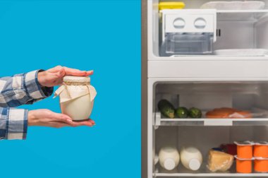 Açık buzdolabının yanında ekşi krema tutan bir kadın ve mavi raflarda taze yiyecek olan dondurucu.