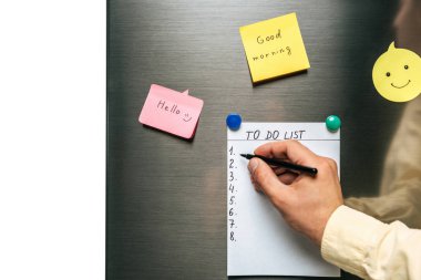 cropped view of man filling in to do list hanging on fridge near wishes on sticky notes isolated on white clipart