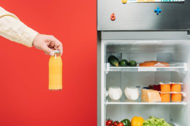 Buzdolabının yanında meyve suyu şişesi tutan adamın kırpılmış görüntüsü ve kırmızı raflarda taze yiyecekler var.