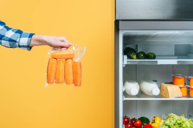 Açık buzdolabının yanında havuç taşıyan, sarı raflarda taze yiyecekler bulunan bir kadının görüntüsü.