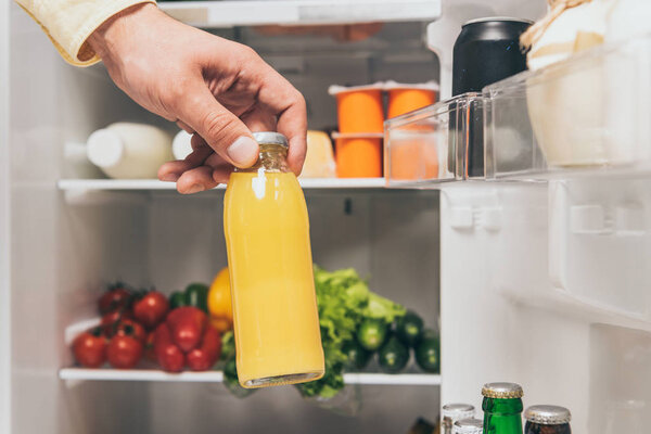 cropped view of man holding bottle of juice near open fridge with fresh food on shelves